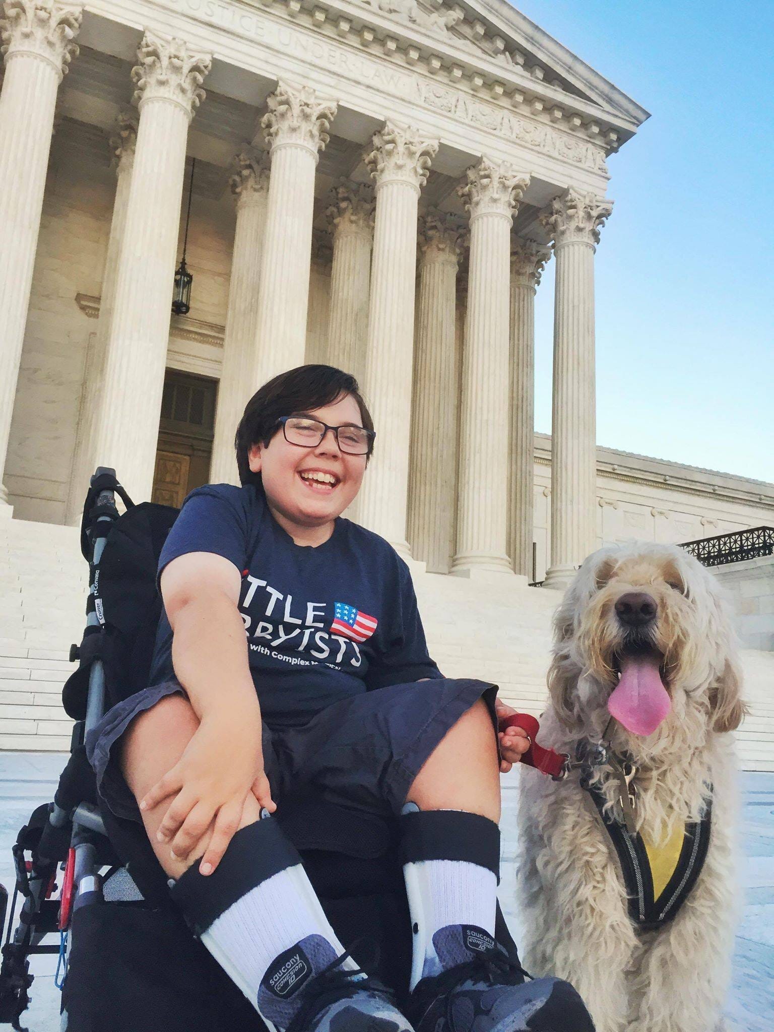 Little Lobbyist Simon Hatcher poses in front of the U.S. Supreme Court steps with his dog. [image description: A light-skinned disabled boy poses in his wheelchair in front of the white marble steps of the U.S. Supreme Court. The Court’s Corinthian columns and can be seen in the background. The boy holds the leash of his dog, a large curly white-haired breed whose long pink tongue is sticking out.]