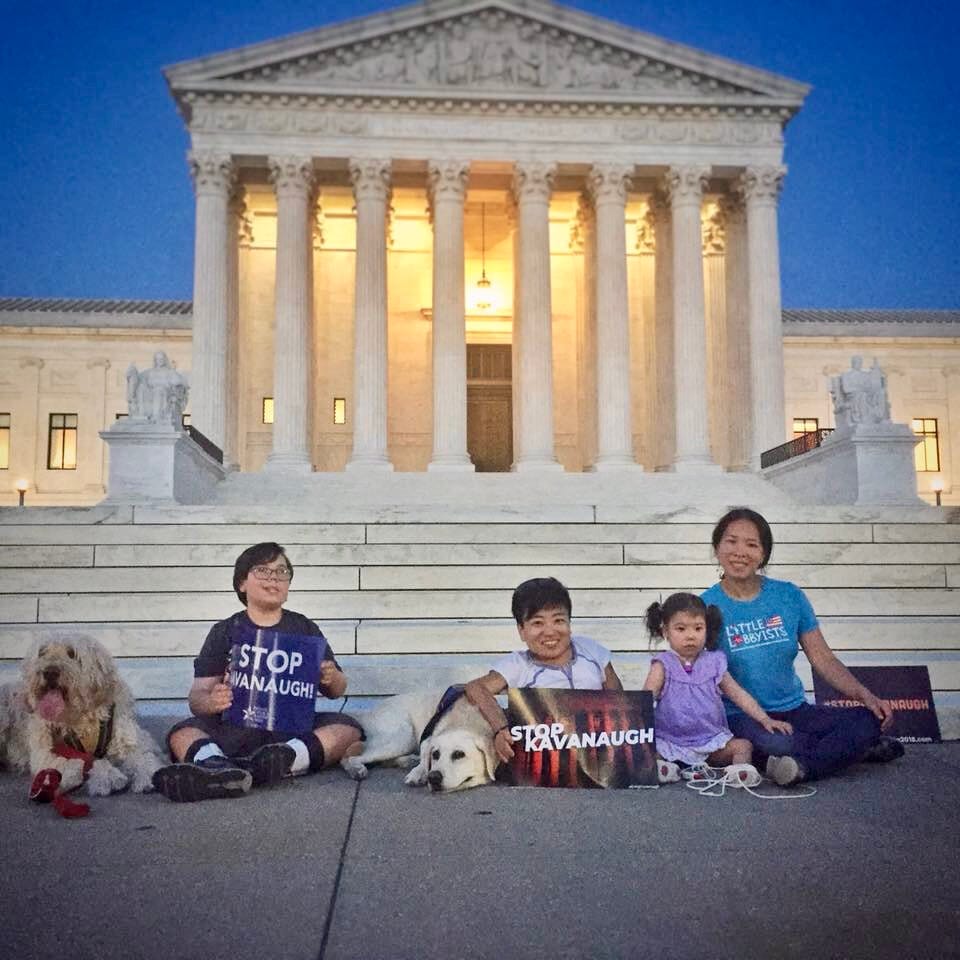 Disability activists Mia Ives-Rublee (center, Director of the Disability Justice Initiative at the Center for American Progress) and Elena Hung (right, Co-Founder of Little Lobbyists), two young disability activists, and two dogs in front of the U.S. Supreme Court. [image description: Left to right are seated on the pavement a large white curly-haired dog, a light-skinned boy with glasses, a large white short-haired dog, a light-tan skinned woman with short black hair, a light tan-skinned young girl with black pony tails, and a light-tan skinned woman with black hair pulled back. In the background is the facade of the U.S. Supreme Court.]