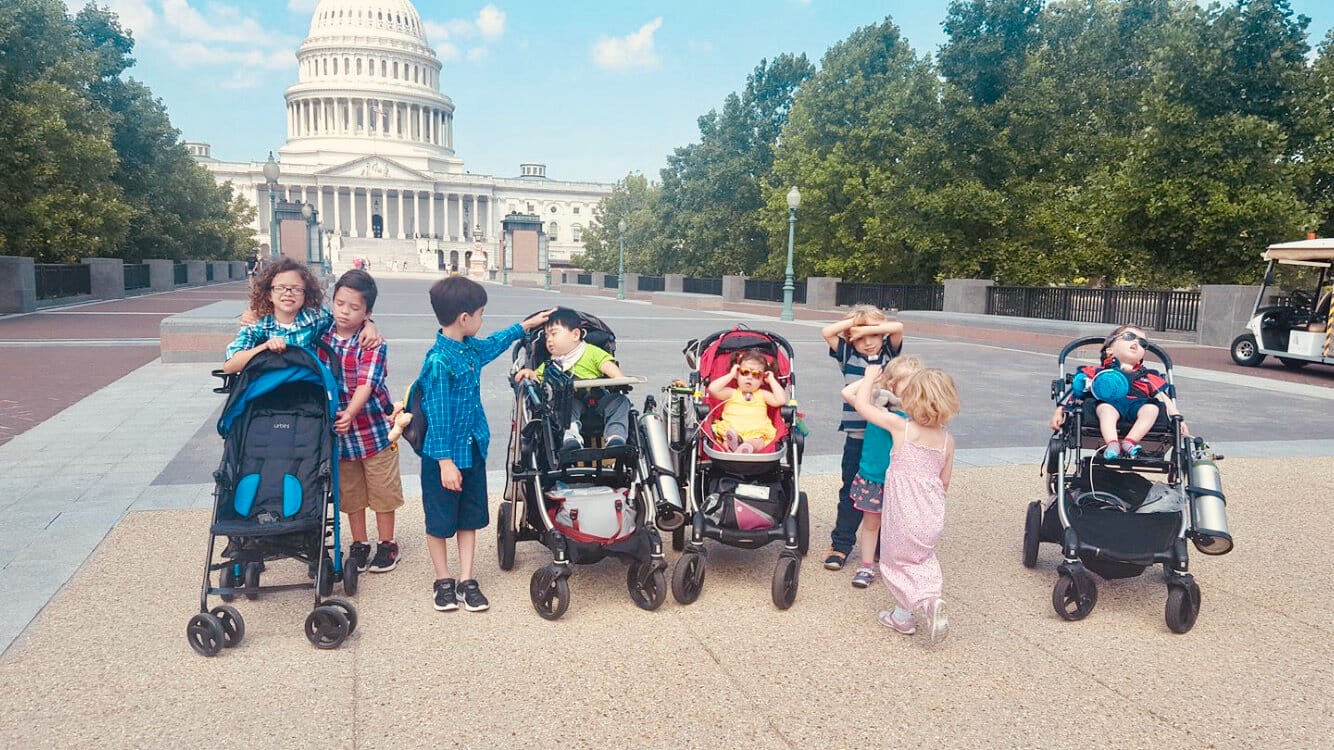 Nine young Little Lobbyists pose on a tree-lined Washington D.C. street with the U.S. Capitol building in the background. [image description: Nine young children with various skin tones pose on a city street with varying shades of pavement (grey, dark red, tan). Some of the children are in adaptive strollers with medical equipment, others stand. They are all goofing off, adorably.]