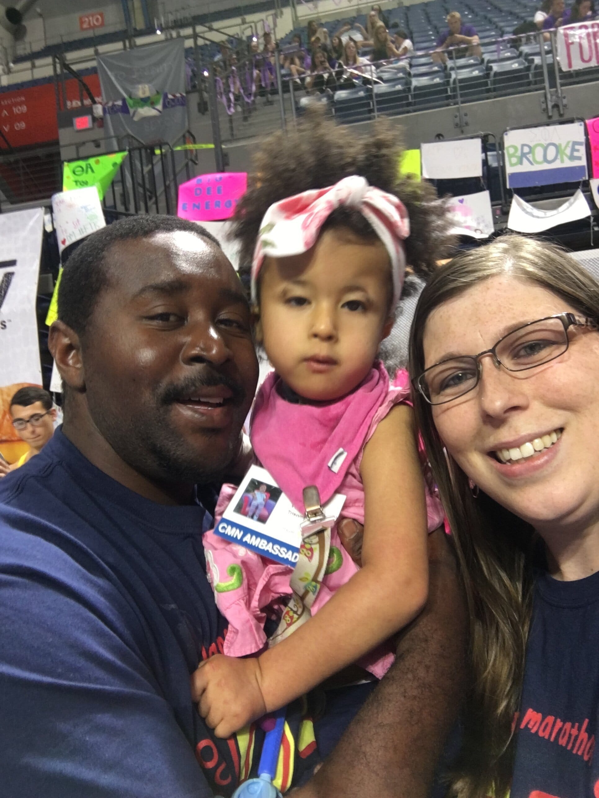 Payton (center) with her dad, Randal (left), and her mom, Kristen (right). [image description: A dark-skinned man in a navy blue shirt holds his daughter, who had tan skin and is wearing a pink dress and a pink and white hair bow. The girl’s mother, a white-skinned woman is to the right with long straight brown hair and glasses.]