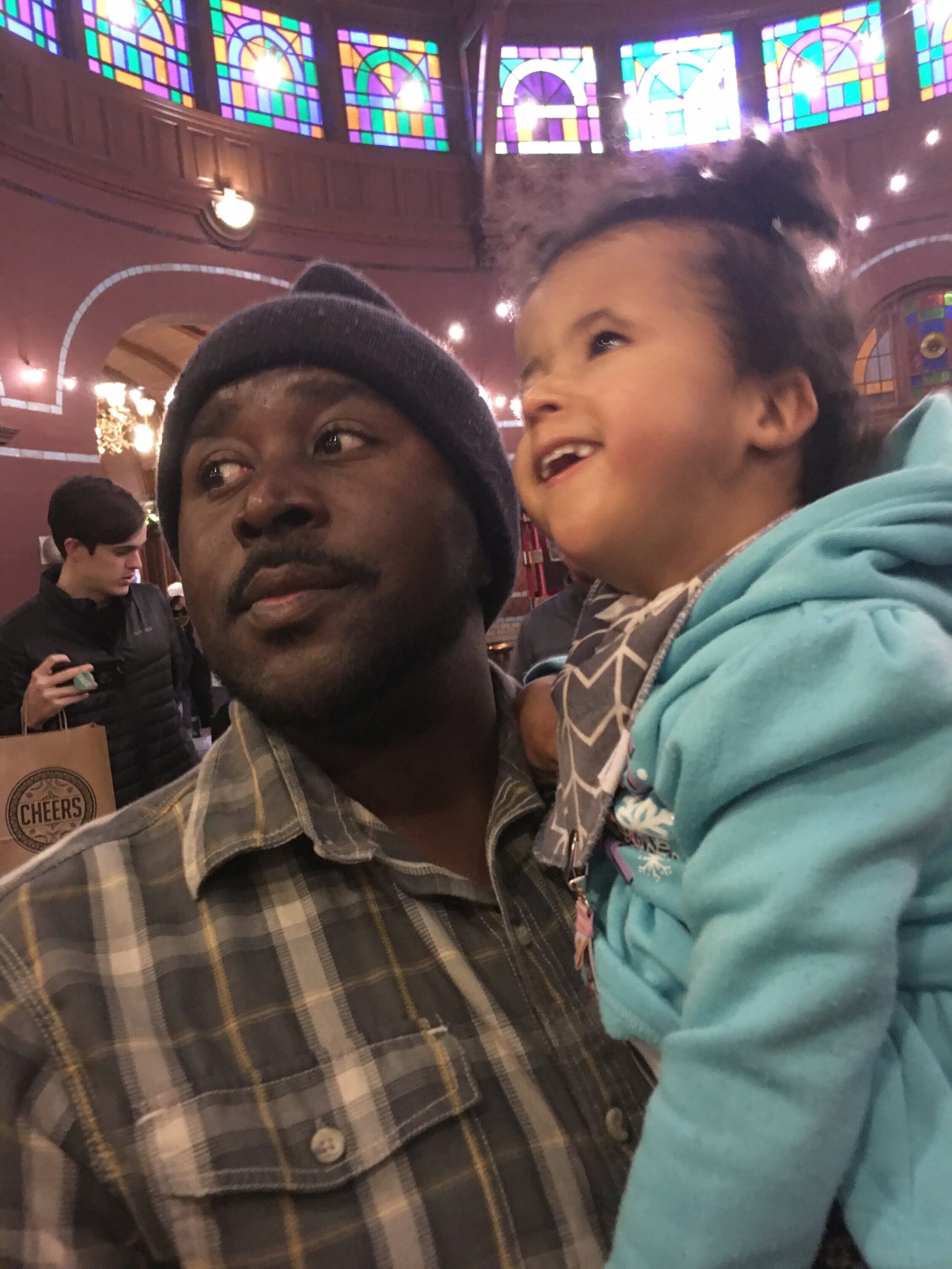 Payton (right) with her dad (left). [image description: A dark-skinned man wearing a knit cap and brown plaid shirt holds his young daughter, who has tan skin and is wearing an aqua jacket. She is looking at bright stained-glass windows.]