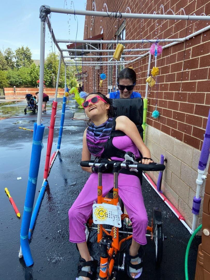 Cailyn out in her community, assisted by a direct support aide. [image description: A young disabled teen wearing pink sunglasses and a bright pink outfit sits on an adapted red bike. An aide assists her moving through a sensory “tunnel” (an open pipework on which various objects are suspended).]