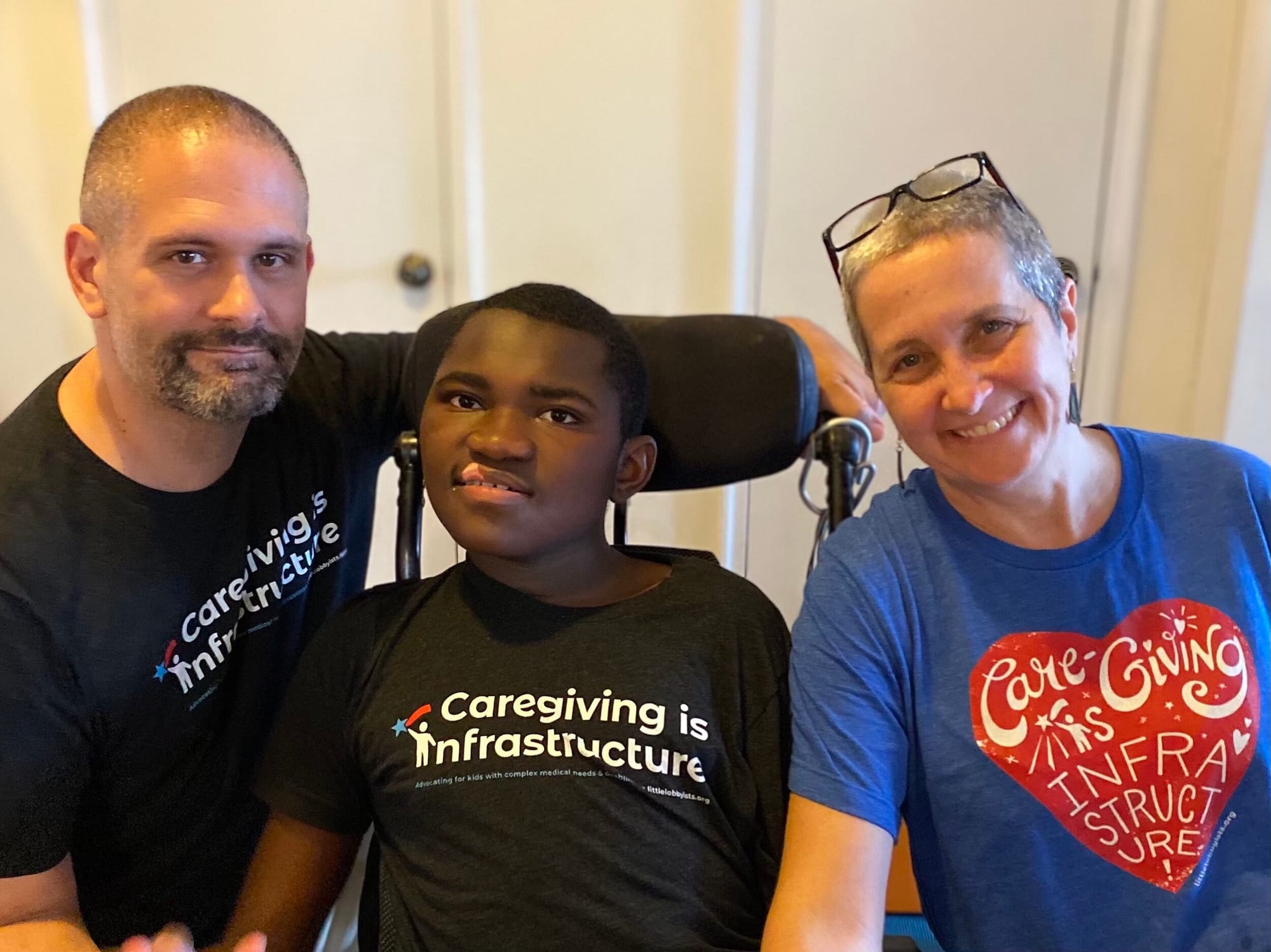 Sandra Stein (r) with her husband Matt (l) and their son Ravi (center) [image description: A white man and white woman pose on either side of a young Black man seated in a wheelchair. All three wear t-shirts with the words, “Caregiving Is Infrastructure.”]