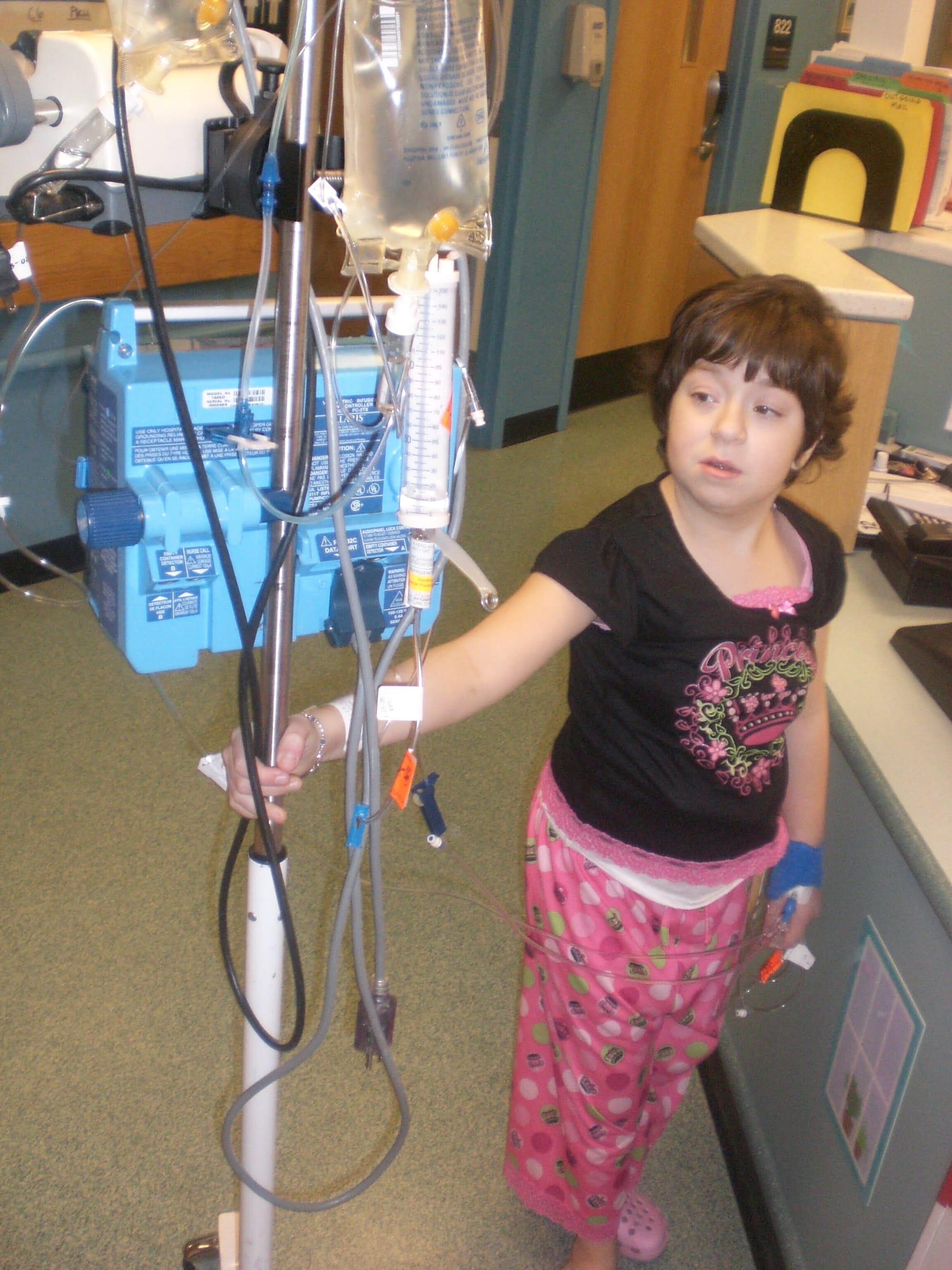 Sierra as a child during a hospitalization. [image description: A young white girl with short black curly hair stands holding onto an IV pole mounted with various medical devices. She wears a black pajama shirt and pink patterned pajama bottoms.]