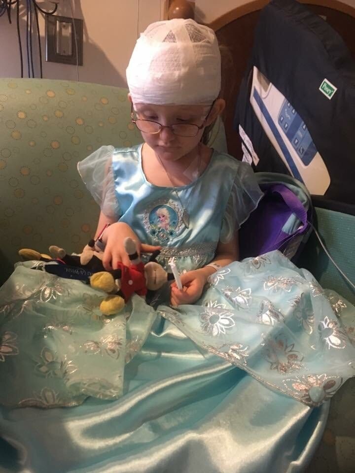 Photograph of Abby, a 6 year old girl wearing an Elsa nightgown, glasses and bandages around her head wrapping up EEG wires while sitting in a chair next to a hospital bed holding Mickey Mouse and Minnie Mouse dolls.