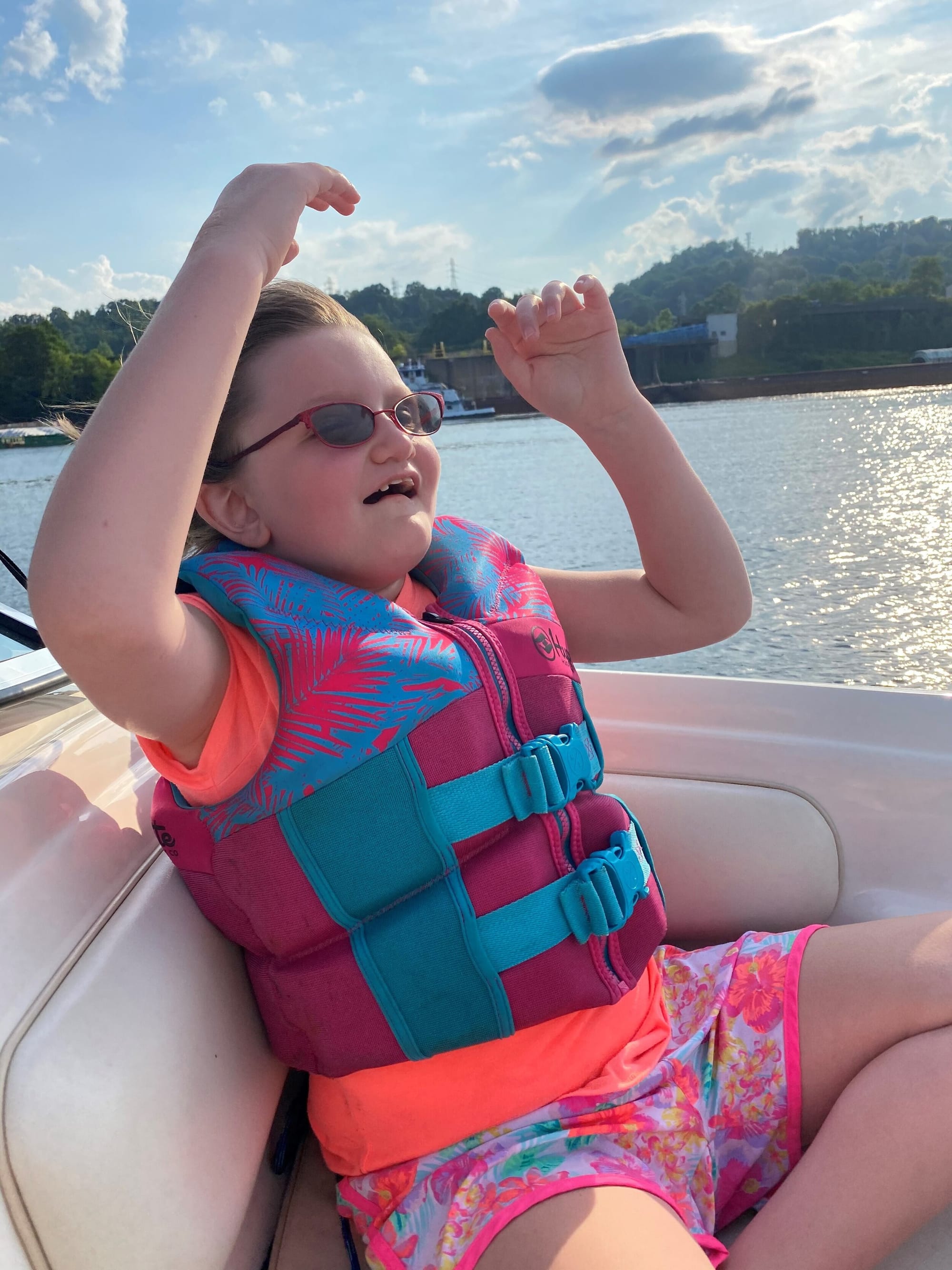 Photograph of Abby, a little girl with brown hair and glasses, now age 11, wearing a pink life vest, seated in the front of a boat on a river, with her hands up in the air and laughing excitedly as though on a roller coaster ride.