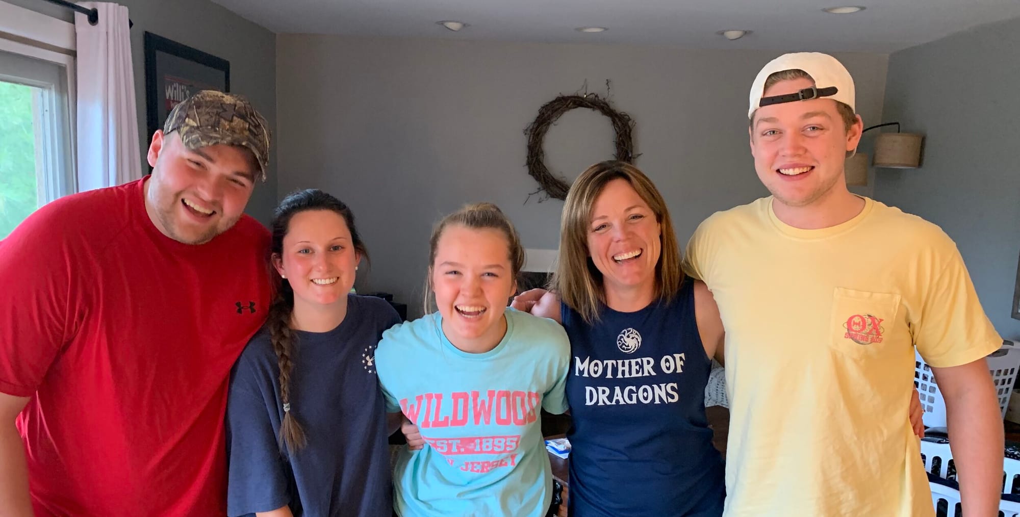 Ande Kolp (third from left) and her children. [image description: A white-appearing family poses in the living room of their home. Five people stand in a line, arms linked. They wear brightly colored t-shirts: 3 women at center, 2 men on either end.]