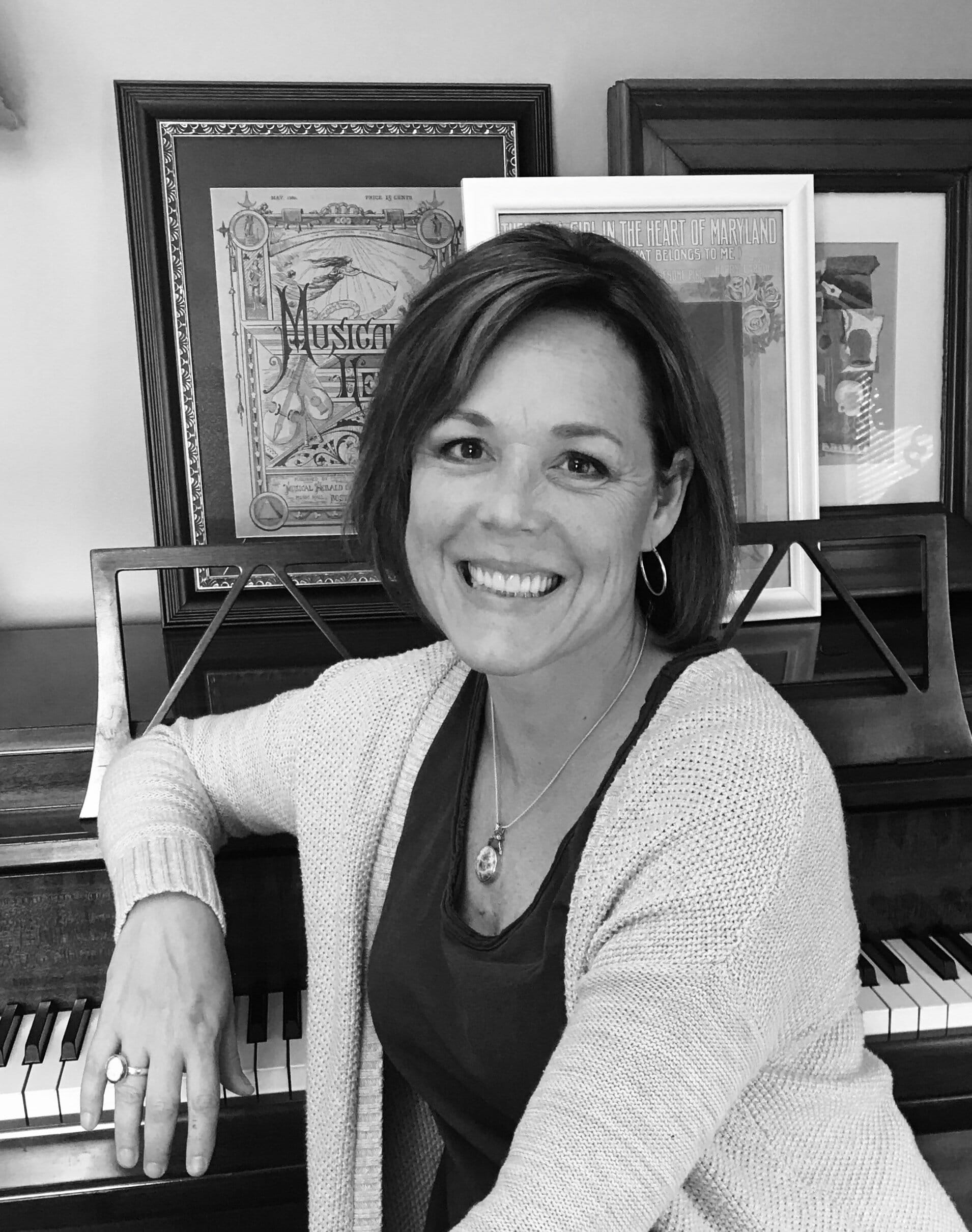 Ande Kolp [image description: A black & white photo of a white-appearing woman with shoulder-length hair, wearing hoop earrings, a light-colored sweater & a dark v-neck top. She poses seated at a piano with framed photos of artwork above the…