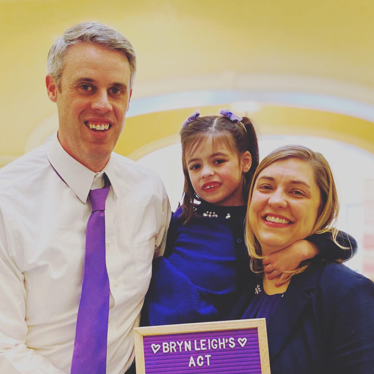 The Shillinger Family. [A father poses in white shirt and purple tie, a mom holds her daughter—both mother and daughter are wearing deep blue clothing.]