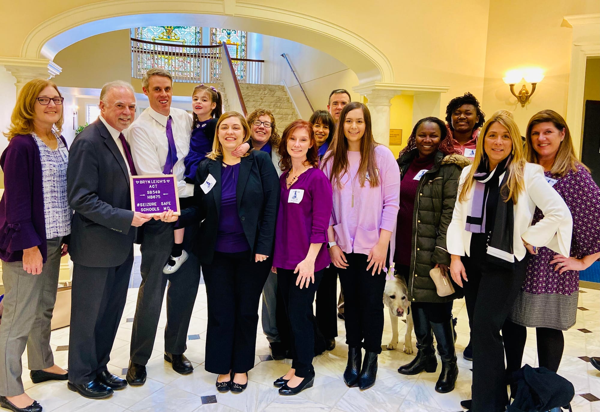 Advocates for Brynleigh’s Act pose in a Maryland General Assembly office building. [image description: Fourteen people wearing shades of lilac, purple and deep blue pose with a Maryland state representative, who holds a sign explaining Brynleigh’s A…