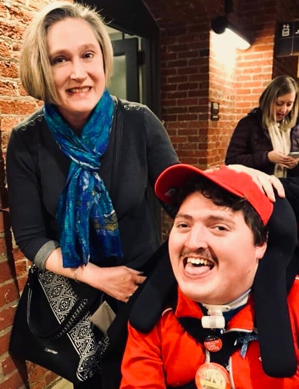 Jeneva &amp; her son Rob [image description: A mom with short blonde hair wears a blue scarf &amp; grey dress. Her son is seated in a wheelchair with an orange shirt &amp; orange baseball cap.]
