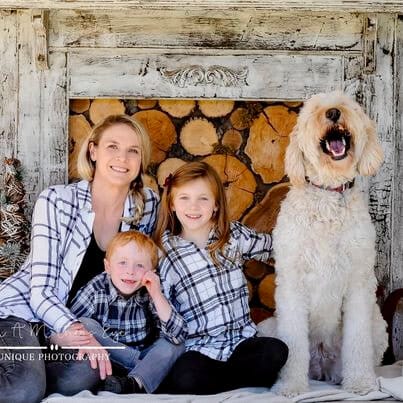 Mallorie and her family. [image description: A family, including a mom, a young boy and a young girl, pose in front of a rustic photographer’s backdrop that includes hewn logs. The family wears coordinating navy blue and white plaid button-down shir…