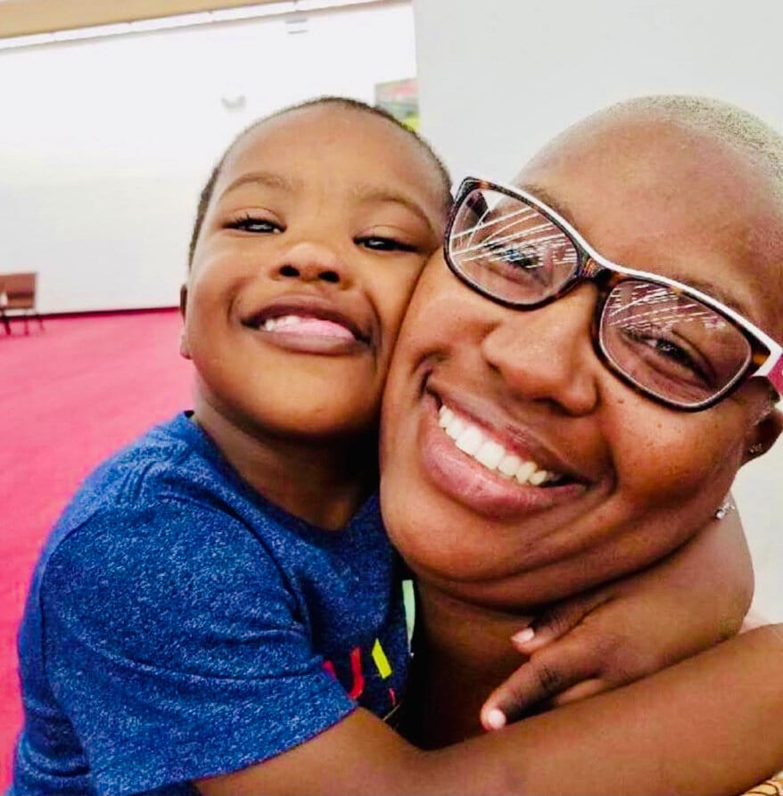 Anian & Meeka [image description: A Black boy with Down Syndrome hugs his mom, who wears glasses and has short light hair.