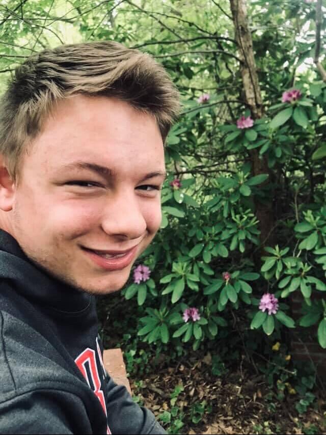 Sam has blonde hair and is wearing a sweatshirt. He is outdoors, looking at the camera and smiling.
