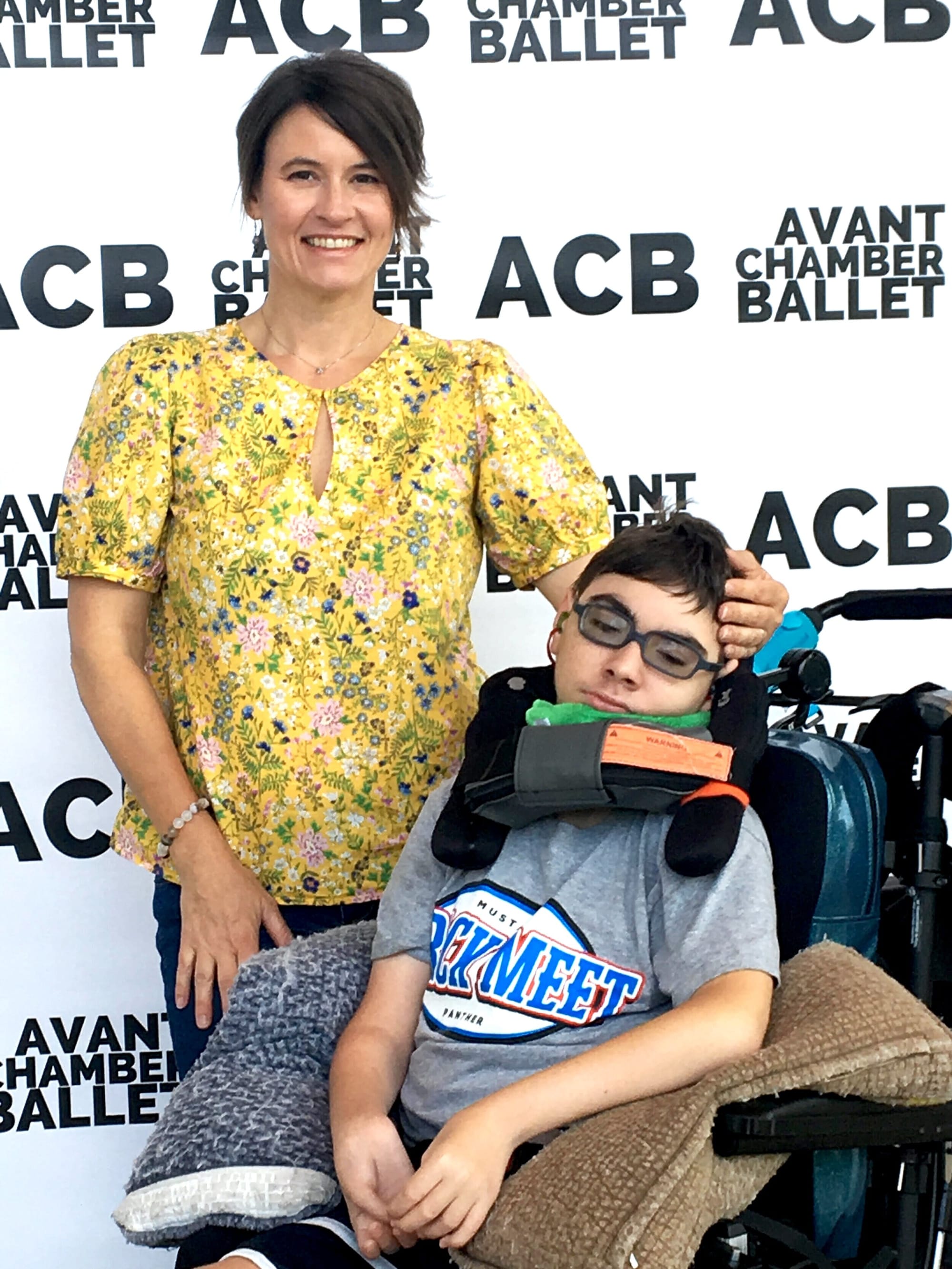 Laura Leeman and her son Vic. [image description: Two people pose in front of a black & white background printed with the words “Avant Chamber Ballet.” A woman with short dark hair & a yellow print shirt stands next to a young man with short…