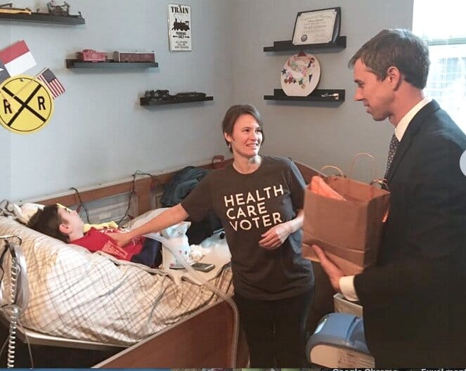 Laura Leeman stands next to Vic (who is in his bed) & speaks with former congressman Beto O’Rourke. [image description: A woman with a “Health Care Voter” t-shirt faces a a man in a suit and tie. They are talking. Next to them, a young disabled …