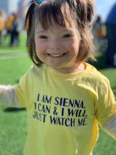 Sienna Striner. She has short brown hair, a big smile, and wears a yellow t-shirt with the words, “I am Sienna. I can & I will. Just watch me.”