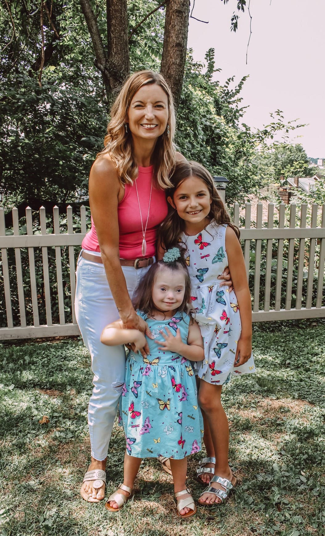Shannon Striner with her daughters Haley and Sienna. Shannon has long blond hair & wears a bright pink sleeveless shirt and white jeans. Haley has long brown hair & wears a sleeveless white dress with a butterfly print. Sienna wears a dress …