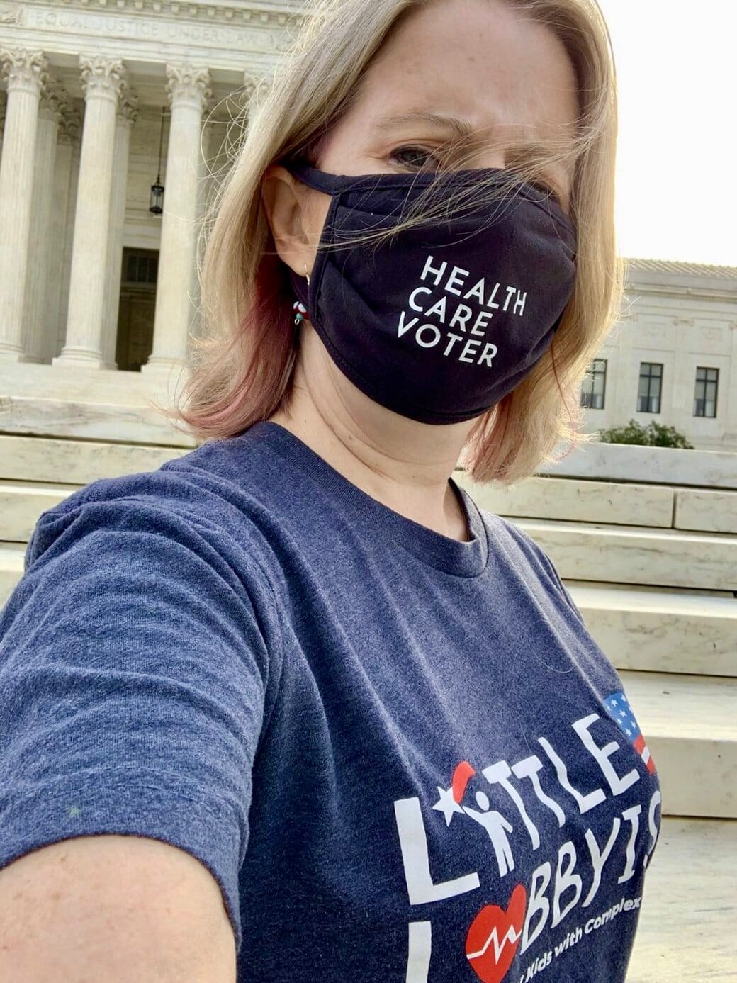 Little Lobbyist Angela Carpenter Gildner at the U.S. Supreme Court after advocating for our families. Angela has blond hair and is wearing a black face mask with the words “Health Care Voter.” She is also wearing a navy blue Little Lobbyists t-shirt…