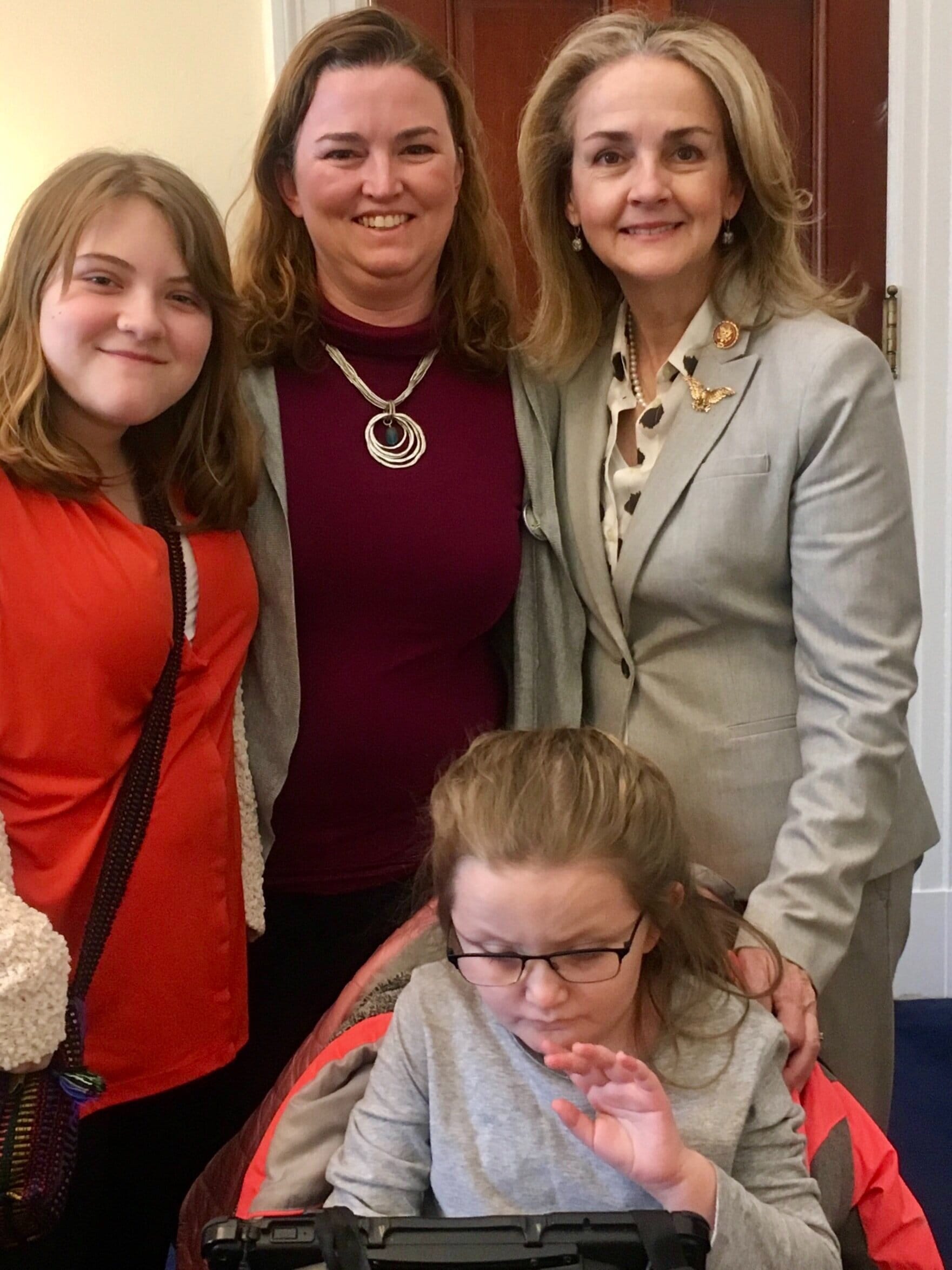 Madeleine Dean standing with Erin & Bridget. Abby is using her speech generating device.