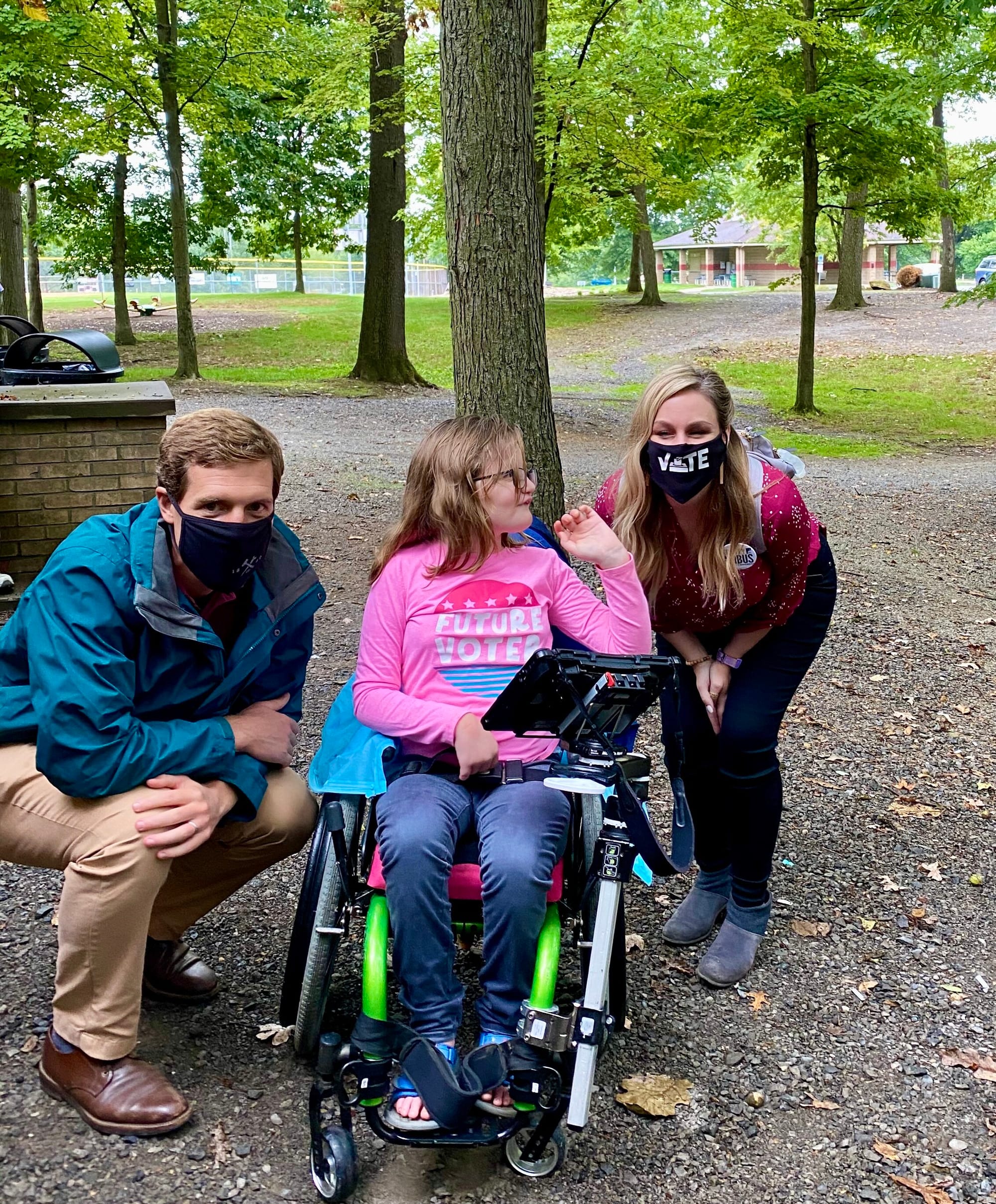 Abby meeting Kristy Gnibus at an outdoor event. Abby is seated in her wheelchair between Conor Lamb, kneeling beside her and wearing a face mask, and Kristy Gnibus, wearing a mask that says “Vote.” Abby is smiling broadly and reaching out to touch G…