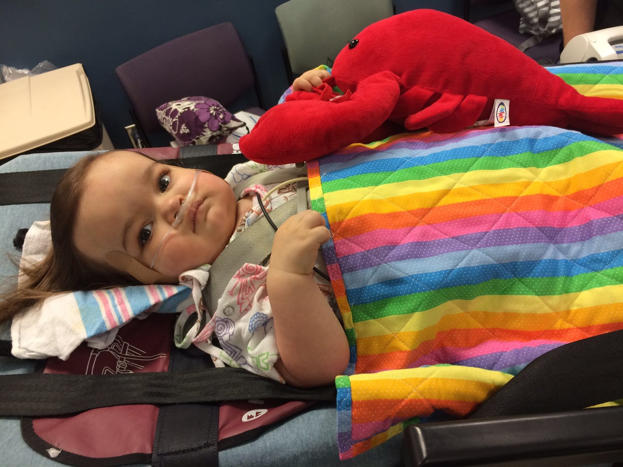 Savannah as an infant. She is strapped to a gurney for medical transport, with a hospital blanket under her head and a rainbow-striped blanket over her torso. A supplemental oxygen cannula has been inserted in both nostrils. She is gripping a bright…