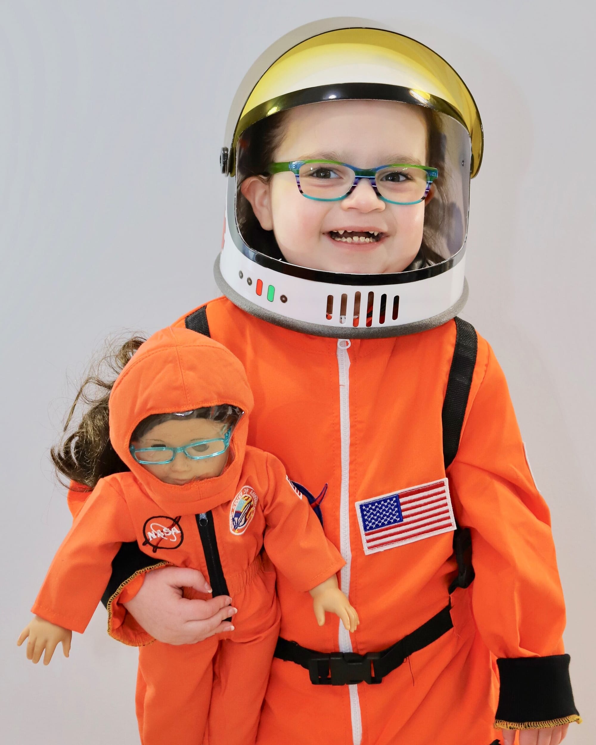Savannah today. She is wearing glasses and smiling. Savannah is dressed in a bright orange NASA space costume, including a space helmet. She is clutching an American Girl doll with her right arm, which is dressed identically.