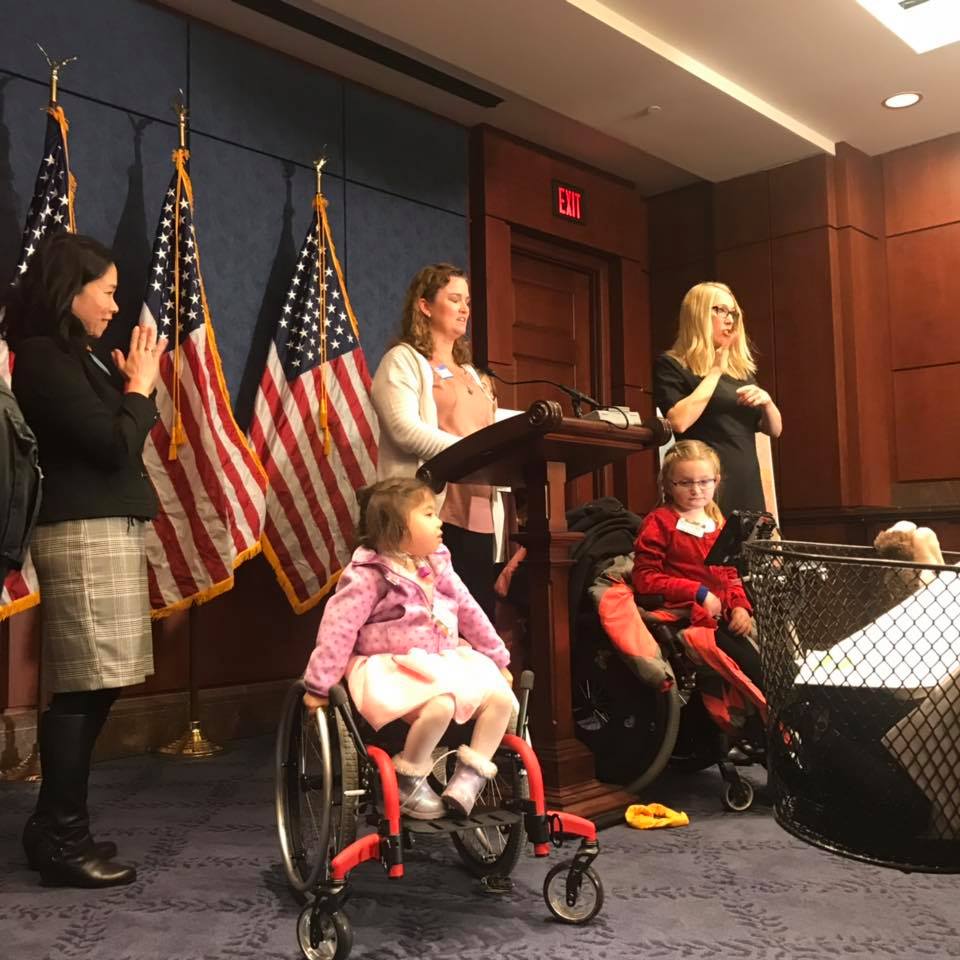 Image Description: Elena Hung, Erin Gabriel, and Little Lobbyists Xiomara and Abby at the podium during the DIA day reintroduction program. They are accompanied by an ASL interpreter.