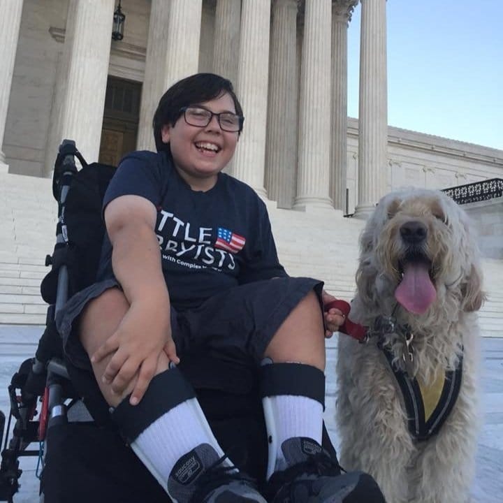 Simon a young teen boy with dark hair and glasses and a big smile. Simon is sitting in a wheelchair, wearing a Little Lobbyists T shirt and holding the leash of his service dog in front of the Supreme Court building