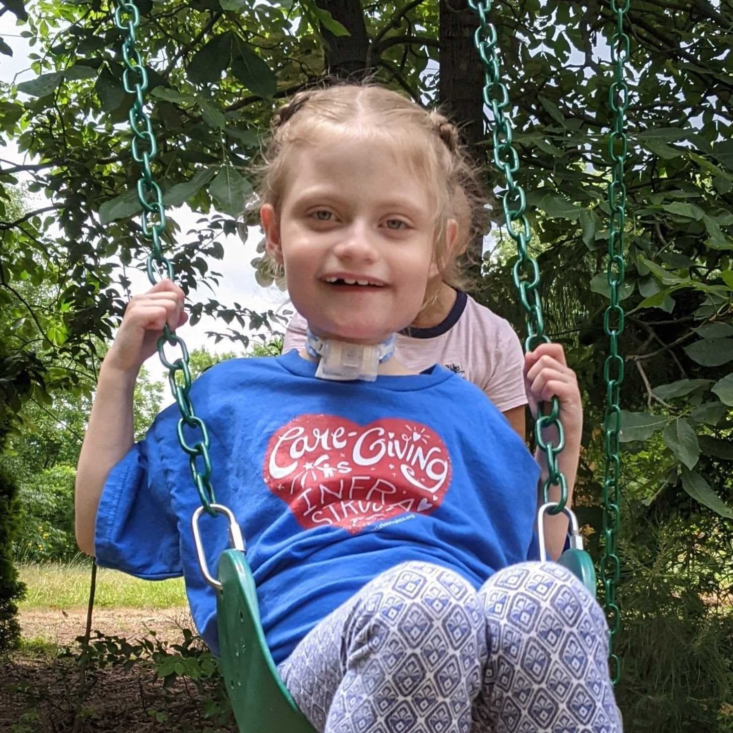 Emma a little girl with blond hair and a tracheostomy wearing a little lobbyists blue shirt swiging on a swing