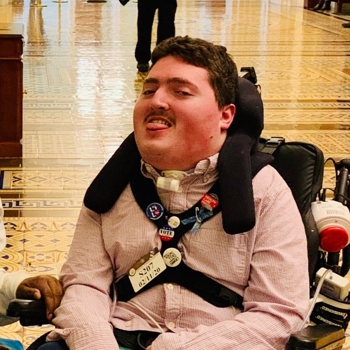 Robert a young man with dark hair, a moustache, and a tracheostpmy is seated in a wheelchair and looking at the camera.