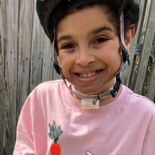 Isaac a smiling young man with a tracheostomy and dark hair wearing a pink shirt and a helmet