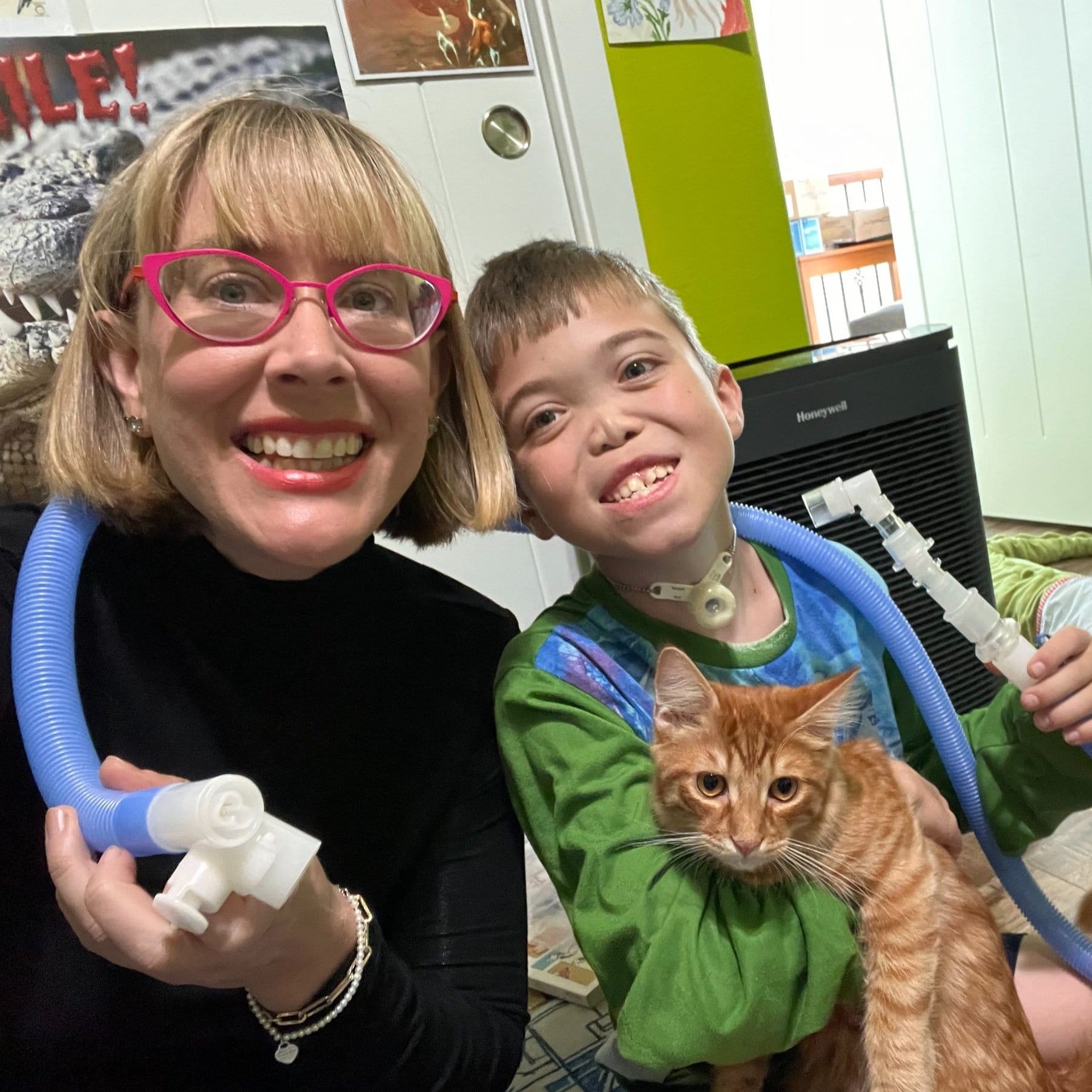 A mom with short blond hair and pink glasses smiles next to her son who is holding an orange cat. The boy looks to be 8-9 years old and has blond hair and a tracheostomy. The mom and the boy are holding a blue ventilator tube festively.