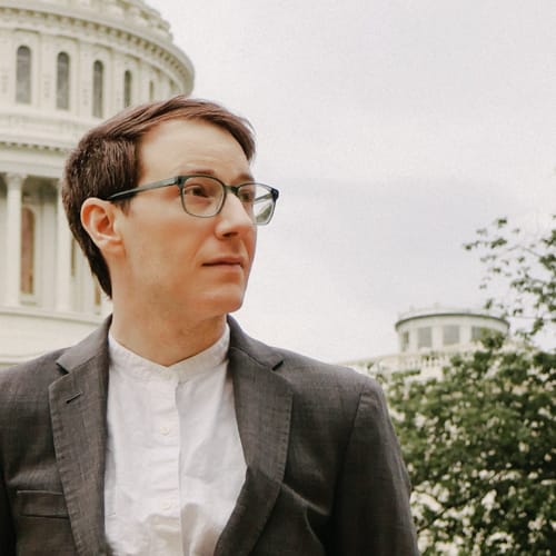 a white non-binary person wearing glasses and a blazer, the U.S. Capitol dome is behind them